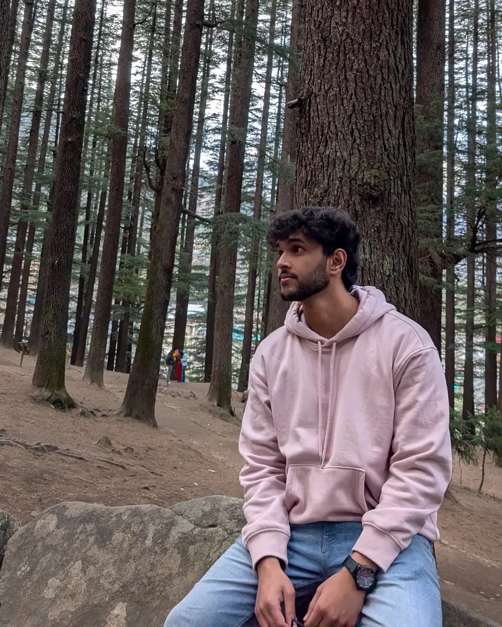 Aditya Sharma sitting on a rock in a pine forest, looking off to the left.